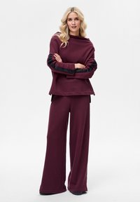 Bordeauxrode trui met zwarte, gestructureerde accenten op de mouwen, gecombineerd met bijpassende wijde broek. Zachte stof, relaxte pasvorm en minimale details.