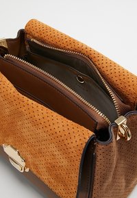 Brązowa torebka z perforowanym pomarańczowym zamszowym klapą, złotymi akcentami i zamykaną na zamek wewnętrzną przegródką. Teksturowane skórzane wykończenia uzupełniają projekt.