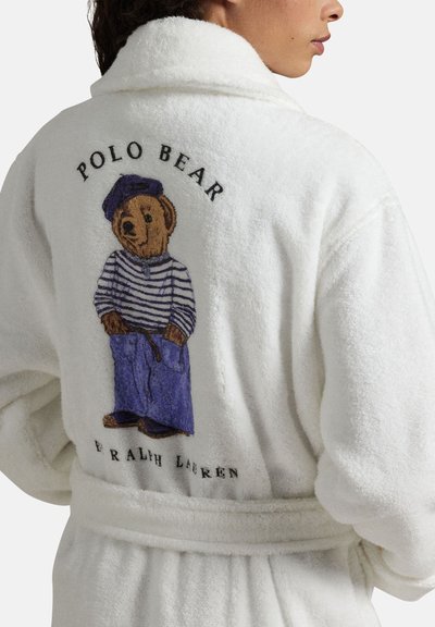Person iført en hvid badekåbe med en brun bjørn i stribet skjorte, lilla bukser og baret broderet på ryggen med teksten "Polo Bear Ralph Lauren."
