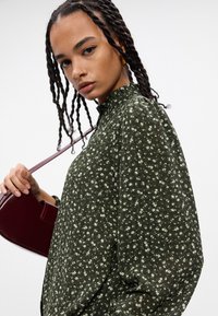 Πράσινη μπλούζα με λουλουδάτο σχέδιο, ψηλό γιακά και φούσκες μανίκια. Το μοντέλο κρατά μια μπορντό τσάντα; τα μαλλιά της είναι χτενισμένα σε μακριές πλεξούδες.