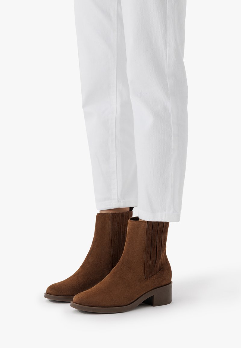 Bruna mockaskinn ankelboots med räfflade sidopaneler och en låg klack, parat med vita rakbenbyxor.