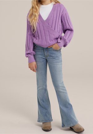 Personne portant un pull en laine lavande sur une chemise blanche, un jean flare bleu clair et des bottes en daim beige, se tenant avec une main dans la poche.