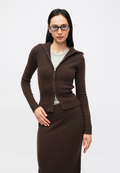 Rjavi pulover na zadrgo z rebrastimi manšetami in robom, v kombinaciji z ujemajočo se rjavo krilo. Pod njim svetlo siva majica. Gladek, pleten tekstura.