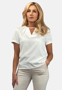 Biała bluzka z krótkim rękawem, z dekoltem w kształcie V i dekoracyjnym złotym akcentem przy dekolcie. Gładka tekstura, luźny krój i subtelne detale drapowania.