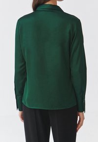 Grön satin långärmad skjorta med krage, med en mjuk yta och knäppta manschetter, stiliserad med en lätt svängd nederkant.