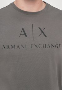 Grå t-skjorte med korte ermer laget av bomull, med en iøynefallende svart "A|X"-logo og teksten "ARMANI EXCHANGE" på forsiden. Glatt tekstur.