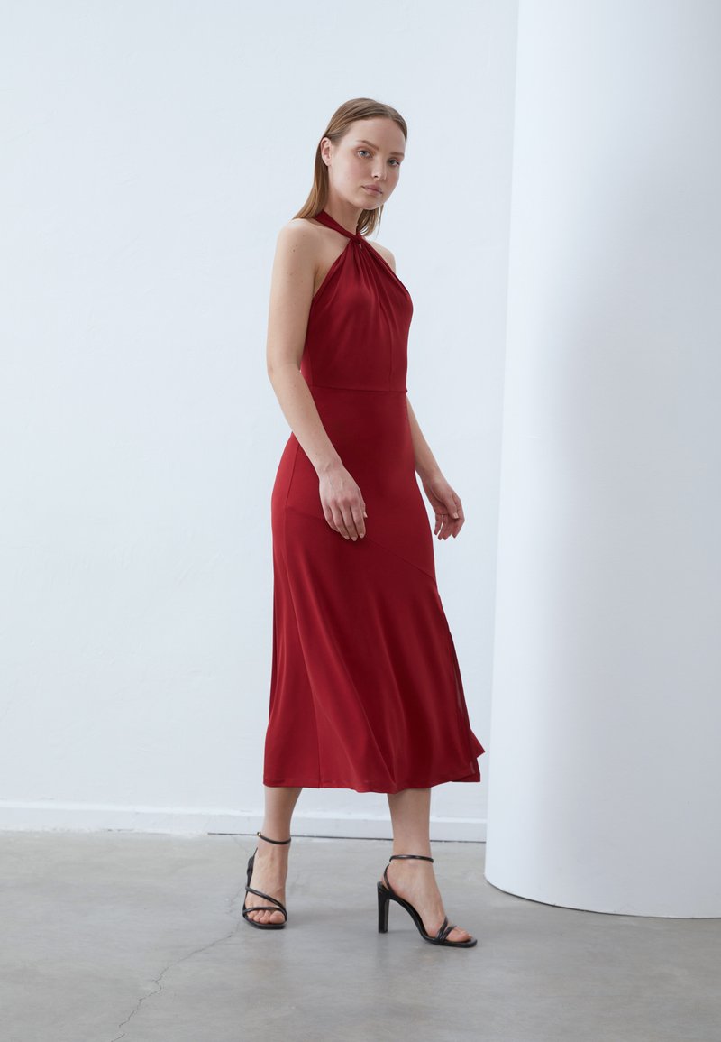 Red, sleeveless dress in a flowing fabric with a halter neck design and ankle-length hem. Paired with black strappy high-heeled sandals.