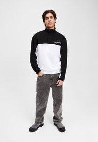 Pullover a mezza zip bianco e nero con maniche lunghe, dettagli con logo, abbinato a jeans grigi baggy e calzature chunky nere. Texture liscia.