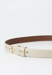 Beige lærrem med gullfarget spenne og logo-detalj, har en glatt tekstur og inkluderer flere justeringshull.