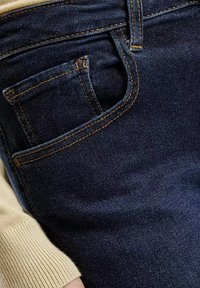 Granatowe dżinsy z przodu z kieszenią, z widocznym szyciem w kontrastowej żółto-pomarańczowej nici, gładka tekstura i dopasowany krój.