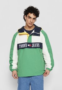 Zaļš, balts un jūras zilas krāsas vēja jaka ar dzeltenu kapuci; ar ievērojamu "TOMMY JEANS" logotipu krūtīs un elastīgām piedurknēm.