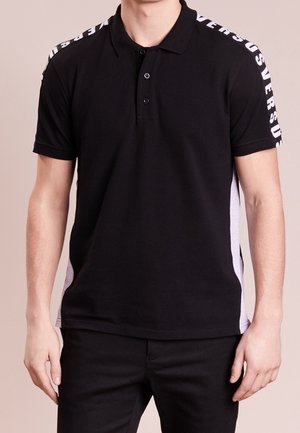 Homme portant un polo noir à manches courtes avec le texte blanc "VERSUS" sur les rayures des épaules et un pantalon noir.