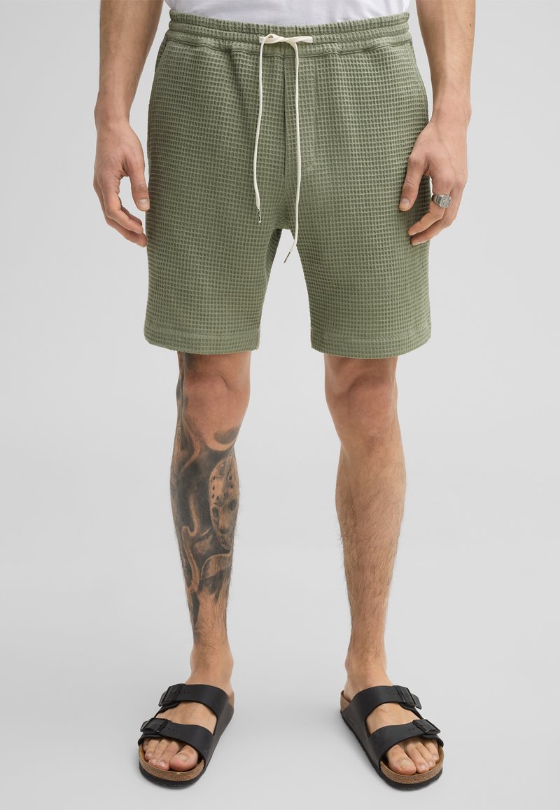 Groene shorts met wafelstructuur, voorzien van een elastische tailleband en een witte trekkoord. Gedragen met zwarte sandalen. Gedetailleerde tatoeage zichtbaar op het linkerschambeen.