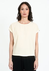 Krēmkrāsas īsroku blūze ar apaļu izgriezumu un atlocītām manšetēm, izgatavota no gludas auduma. Valkāta kopā ar melniem biksēm.