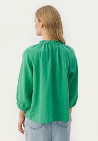 Grüne Leinenbluse mit einem Rüschenausschnitt, langen, luftigen Ärmeln und einer Rückenfalte. Sie hat eine lockere Passform und eine weiche Textur.