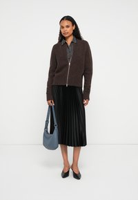 Γυναίκα που φοράει καφέ φούτερ με φερμουάρ πάνω από ριγέ μπλουζίτσα, μαύρη πλισέ midi φούστα, μαύρα παπούτσια, κρατώντας μπλε τσάντα ώμου, στέκεται μπροστά σε λευκό φόντο.