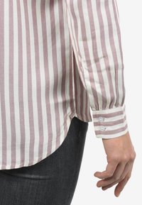 Camicia a maniche lunghe a righe, rosa chiaro e bianco, con polsini a bottoni e orlo curvo. Tessuto testurizzato con cuciture visibili.