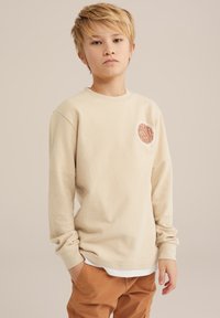 Chemise à manches longues en maille gaufrée beige avec un logo rond sur la poitrine, associée à un pantalon cargo brun. Présente un design décontracté et une coupe ample.