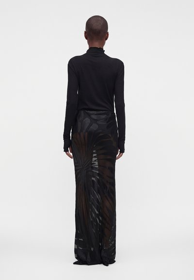 Personne debout de dos à la caméra, portant un haut noir à manches longues et une longue jupe noire transparente à motif abstrait tourbillonnant.