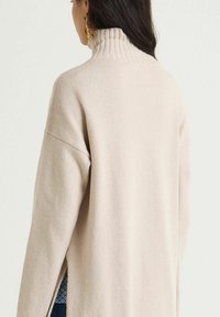 Pull beige avec col montant côtelé, texture douce et épaules tombantes. Fentes latérales pour plus de confort, dévoilant une couche à motif en dessous.