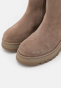 Suede ankle boots in light brown with a rounded toe, thick rubber sole, and contrasting white stitching along the edge. Smooth texture.