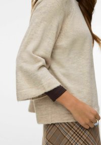 Femme portant un pull en maille beige à manches larges sur un chemisier à manches longues foncé et une jupe à carreaux marron, avec une bague argentée à la main droite.