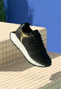 Baskets noires et dorées avec semelle blanche tachetée, lacets noirs et texte "REPLAY 1981", exposées sur des marches carrelées blanches contre un mur bleu.