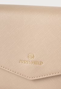 Μπεζ υφασμάτινη πτυχωτή τσάντα με κλείσιμο φάκελου, με χρυσό λογότυπο Anna Field και στρογγυλό έμβλημα στο κέντρο κάτω από το ράψιμο της άκρης.