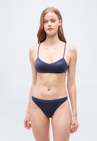 Γυναίκα με μακριά ξανθά μαλλιά που φοράει μια σκούρα μπλε, λαμπερή μπικίνι με λεπτές τιράντες, στέκεται μπροστά σε ένα απλό ανοιχτό γκρι φόντο.