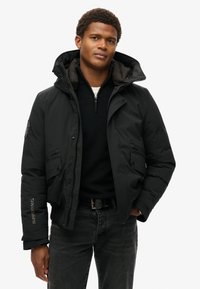 Sort puff jakke med hætte, der har lynlås og to forlommer. Udsmykket med et logo på ærmet, båret over en mørk sweater.