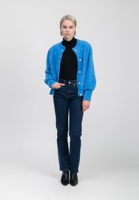 Eine Frau steht in einem blauen, flauschigen Cardigan mit Perlenknöpfen, einem schwarzen Rollkragenpullover, dunklen Jeans und schwarzen Stiefeletten vor einem schlichten Hintergrund.