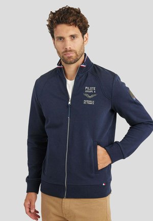 Homme aux cheveux bouclés portant une veste zippée bleu marine avec l'emblème "Pilote Groupe A Patrouille de France", pantalon beige, debout devant un fond gris.