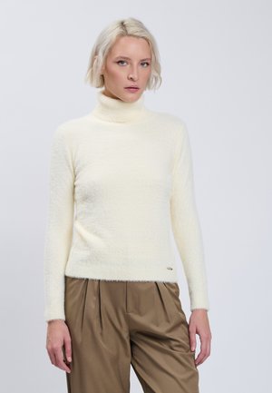 Jonge vrouw met kort blond haar, gekleed in een witte pluizige coltrui en bruine geplooide broek, staand tegen een effen achtergrond.