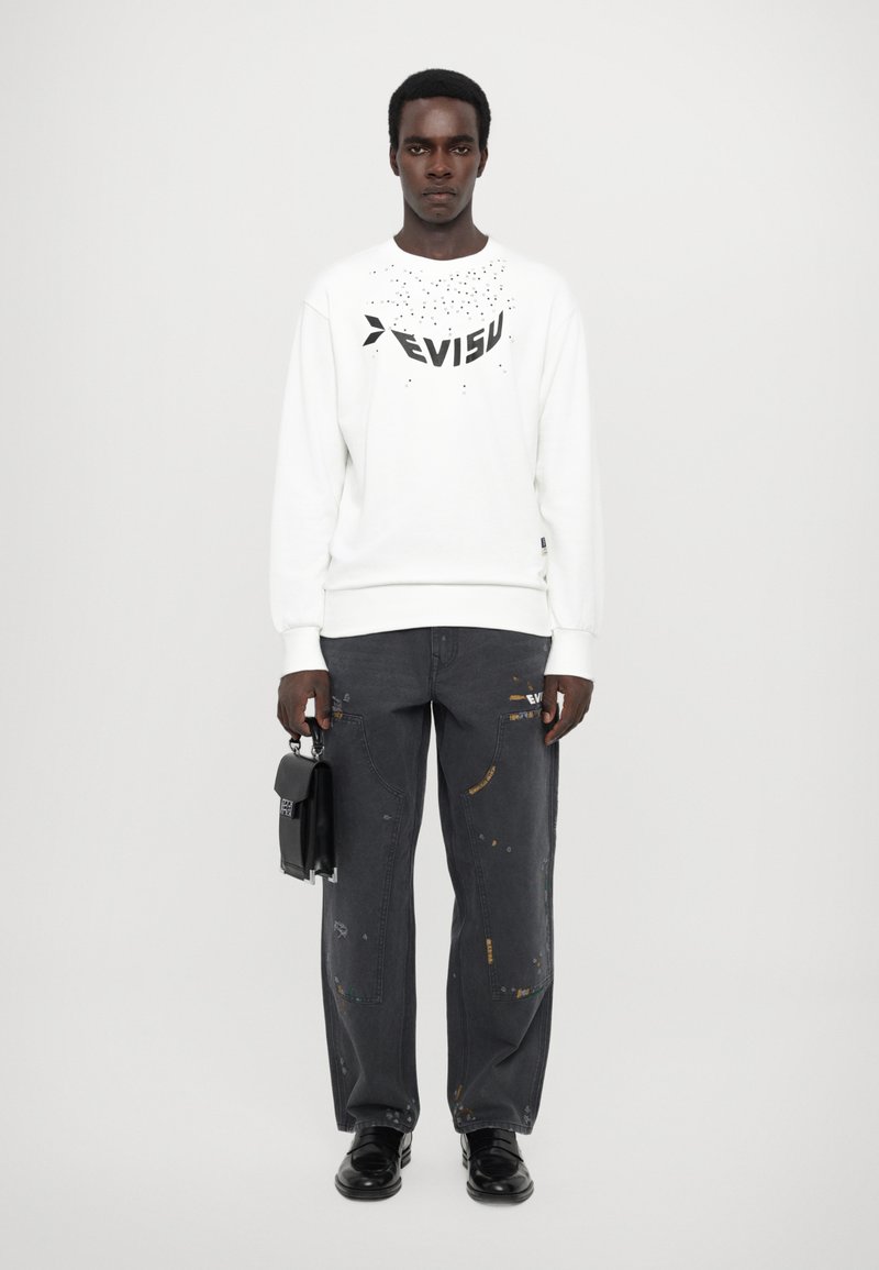 Vit sweatshirt med svart text, kombinerad med lösa grå jeans med färgstänk och en svart väska. Svarta skor fullbordar looken.