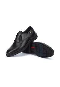 Chaussures de ville pour hommes en cuir noir avec lacets, une chaussure debout et l'autre montrant une semelle en caoutchouc motifs avec un logo rouge.
