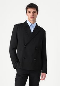 Schwarz, doppelt geschichtete Jacke aus strukturiertem Stoff, ausgestattet mit zwei seitlichen Taschen und schwarzen Knöpfen, getragen über einem hellblauen Hemd.
