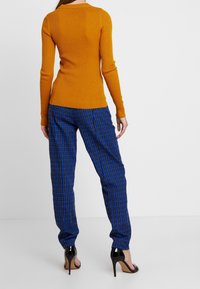 Ribbad senapsgul långärmad topp tillsammans med blå rutig byxa. Byxorna har svarta och gula accentfärger. Modellen bär svarta högklackade skor.