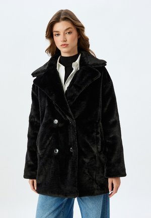 Cappotto in pelliccia sintetica nera con colletto a punte, design doppiopetto, grandi bottoni e tasche laterali, indossato sopra una camicia bianca e un dolcevita nero.