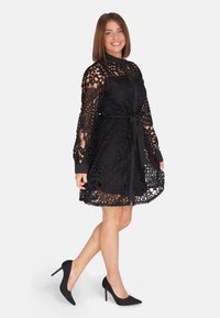 Abito in pizzo nero con bottone frontale, maniche lunghe trasparenti e vita con cintura. Presenta intricati motivi ritagliati e orlo scallop.