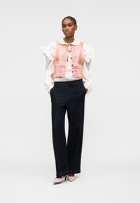 Femme portant un chemisier blanc à manches volantées, un gilet rose à motifs, un pantalon noir ample et des talons blancs pointus, debout les mains dans les poches.