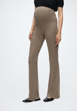 Personne enceinte portant un pantalon de maternité côtelé beige et des chaussures plates noires sur un fond gris clair, les mains reposant sur les côtés.