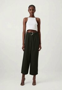 Haut halter blanc court, pantalon noir taille haute avec une boucle de ceinture circulaire en argent, et des talons noirs à lanières. Texture de tissu lisse.