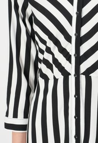 Robe chemise rayée noir et blanc avec un devant boutonné, des manches longues, un buste structuré et une taille ajustée ornée de motifs verticaux.