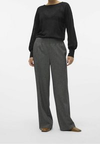 Person klädd i en svart långärmad texturiserad topp, lösa grå högmidjade byxor och bruna spetsiga skor mot en vit bakgrund.