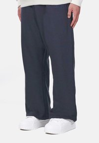 Navy blue housut löysällä mallilla, joissa on teksturoitu kangas ja brodeerattu logo vyötäröllä, yhdistetty valkoisiin tennareihin.