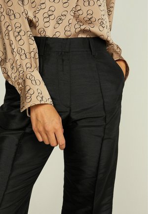 Pantalon noir en satin avec une texture lisse et des détails de plis, associé à un haut beige à motifs avec des cercles abstraits noirs.
