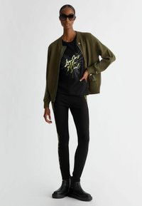 Giacca bomber a coste verde oliva sopra una maglietta grafica nera, abbinata a leggings neri con parte laterale a motivo giallo. Stivaletti neri alla caviglia.