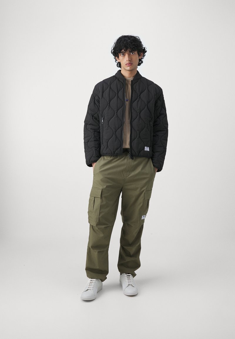 Czarna pikowana kurtka z kołnierzem stojącym, beżowa bluzka pod spodem, oliwkowe spodnie cargo z kieszeniami i białe trampki.