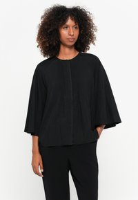 Blusa preta com mangas largas, tecido texturizado e fecho de botões decorativos na parte da frente. Design leve e de corte solto.