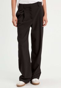 Pantaloni neri a gamba larga realizzati in tessuto liscio, con pieghe frontali, tasche laterali e una cintura su misura. Indossati con scarpe di colore chiaro.